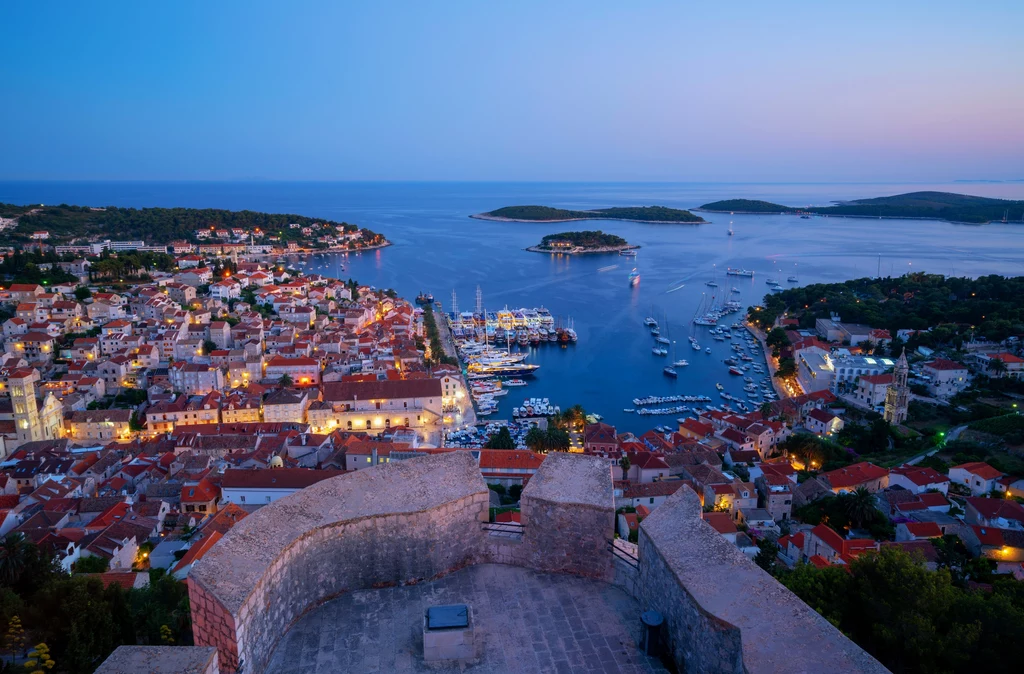 Hvar, Chorwacja Wieczorny widok z perspektywy wzgórza na nadmorskie miasto z licznymi czerwonymi dachami, mariną pełną jachtów i łodzi oraz spokojną zatokę otoczoną wyspami, wszystko oświetlone ciepłymi światłami miasta.