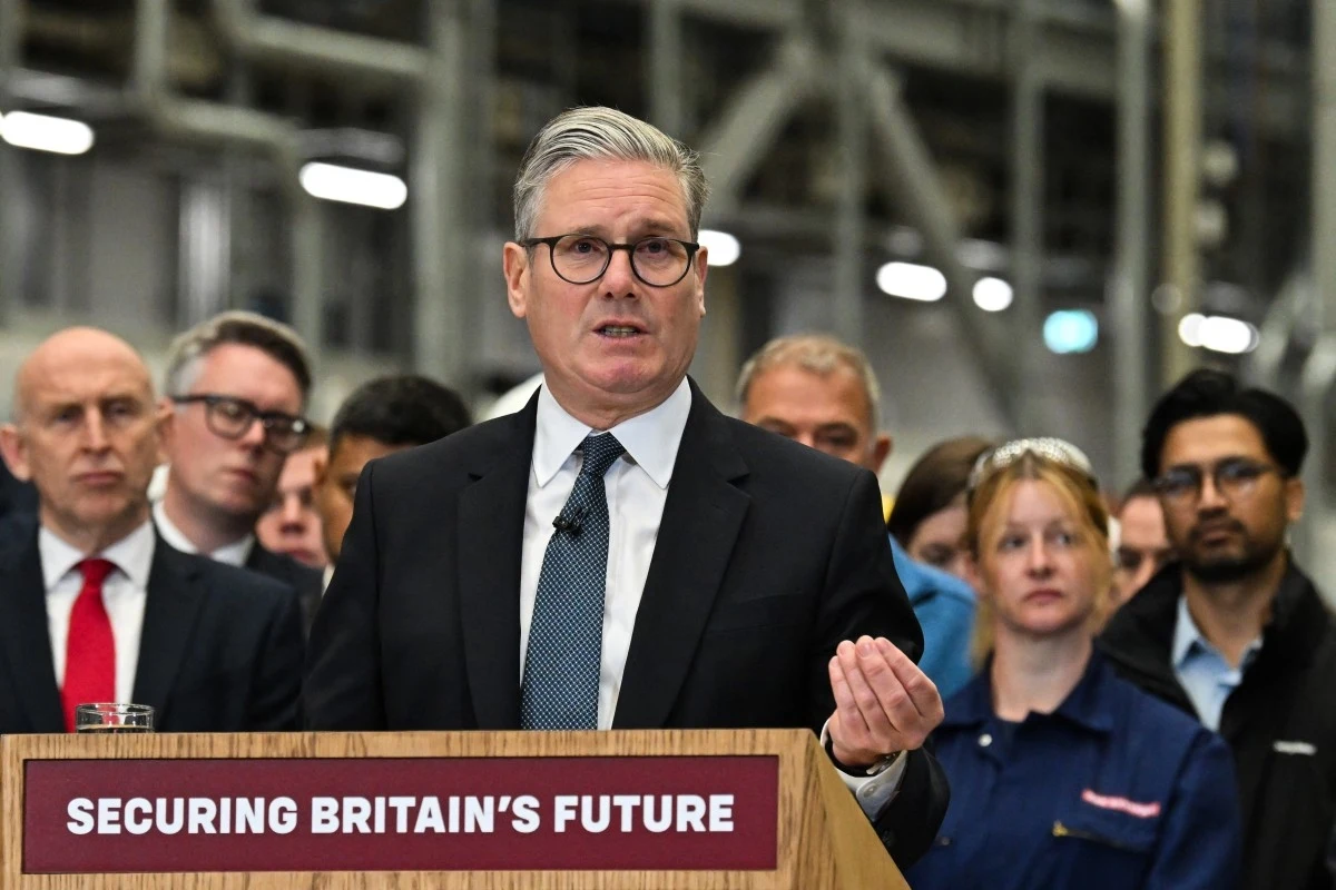 Premier Wielkiej Brytanii Keir Starmer Mężczyzna w garniturze przemawia przy mównicy z napisem 'Securing Britain's Future', za nim stoi grupa poważnych osób w formalnych oraz roboczych ubraniach, tło stanowi industrialne wnętrze.