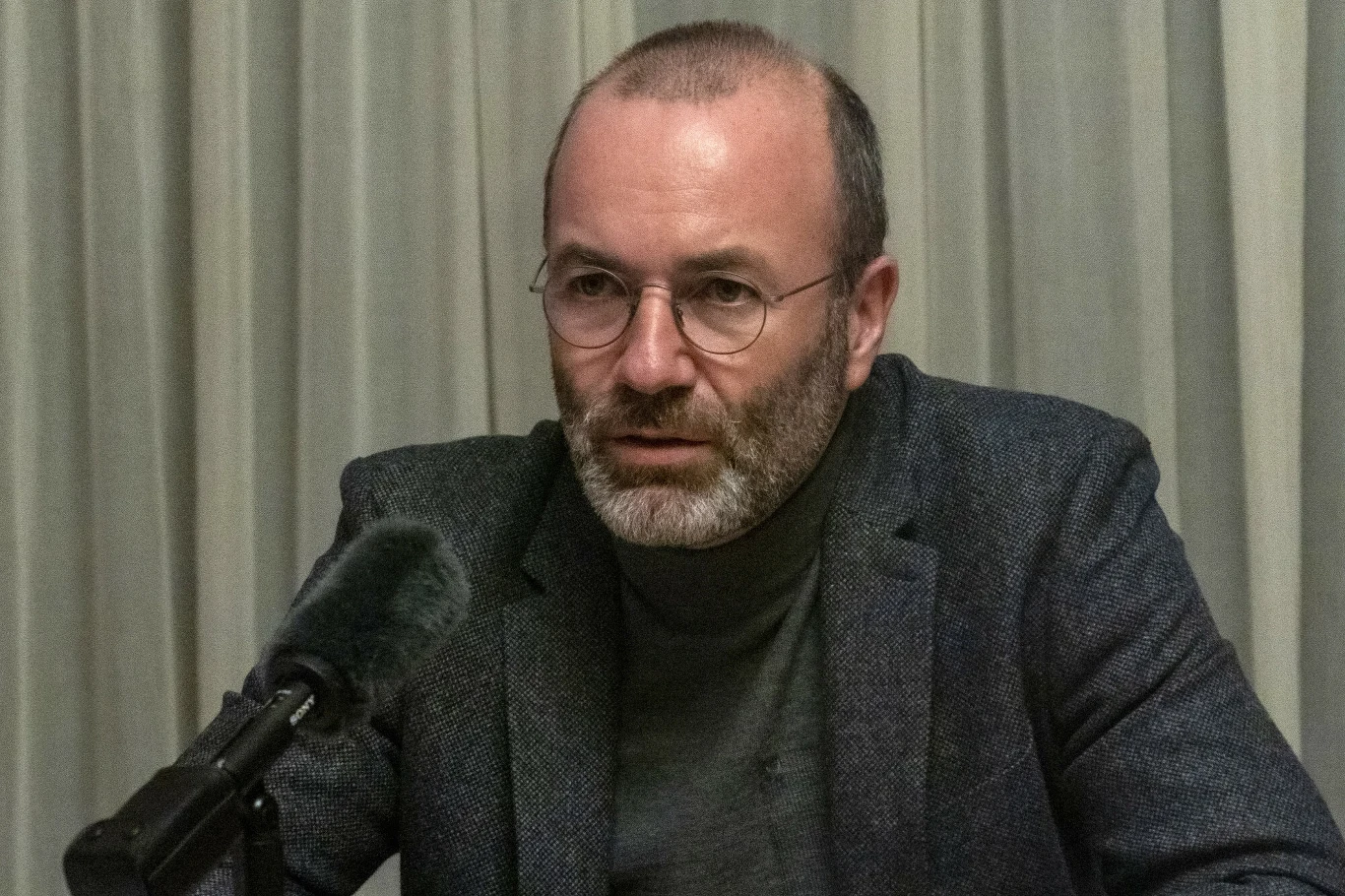 Wybory w Polsce. Manfred Weber reaguje Mężczyzna w okularach o krótkich, siwych włosach i brodzie siedzi przy stole za mikrofonem, ubrany w ciemny golf i marynarkę, w tle jasna zasłona.