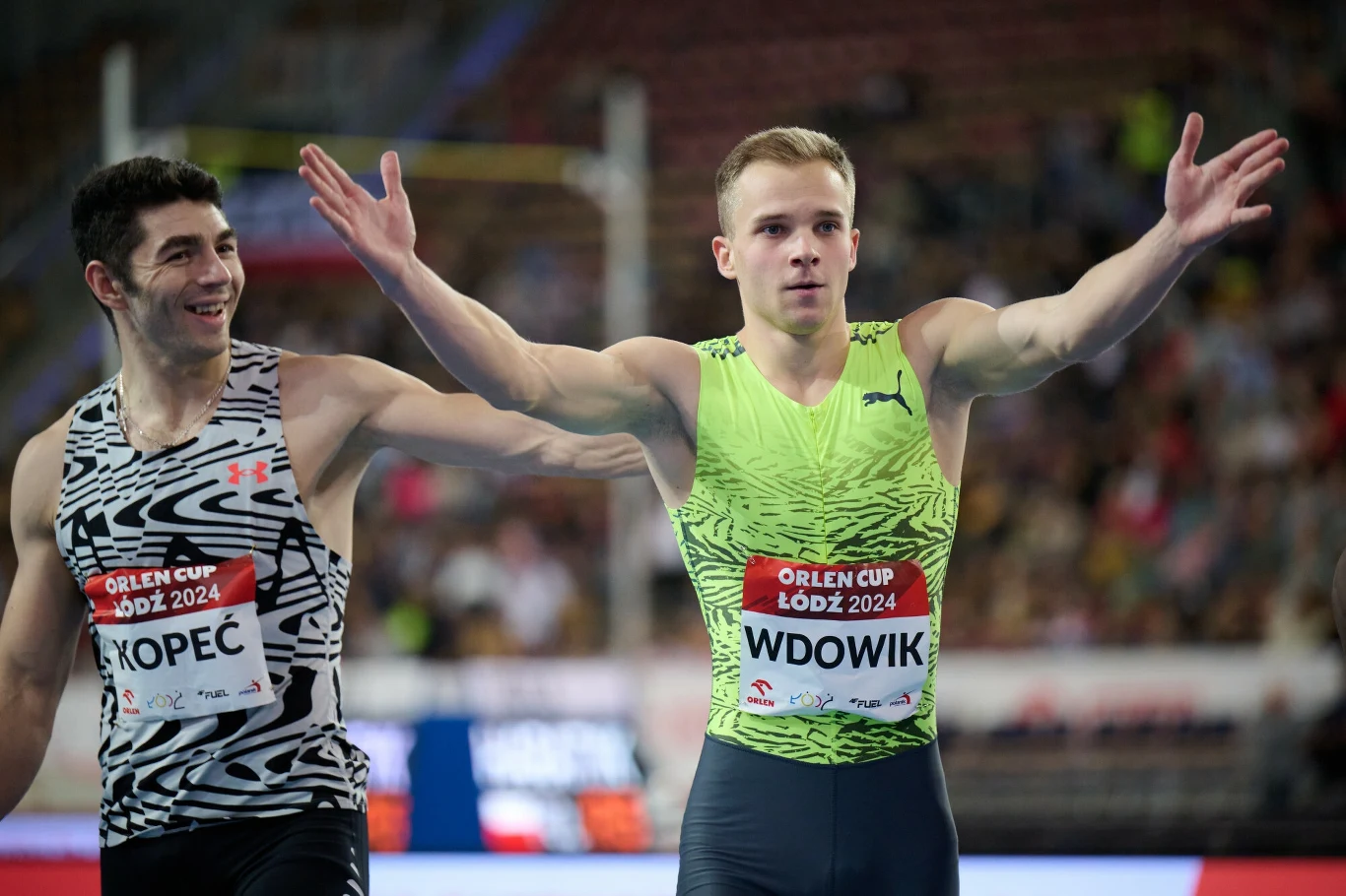 Dwóch lekkoatletów tuż po zakończonym biegu halowym, jeden z zawodników z szeroko rozłożonymi ramionami i tabliczką z napisem Orlen Cup Łódź 2024, trybuny w tle wypełnione kibicami.