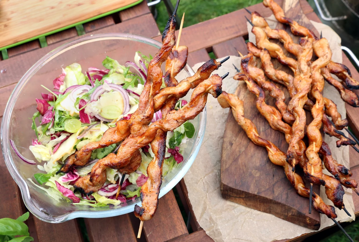 Sałatka z oscypkiem i winogronami Drewniany stół ogrodowy, na którym stoi miska z kolorową sałatką warzywną oraz podłużne grillowane szaszłyki z mięsem poukładane na desce i misce.