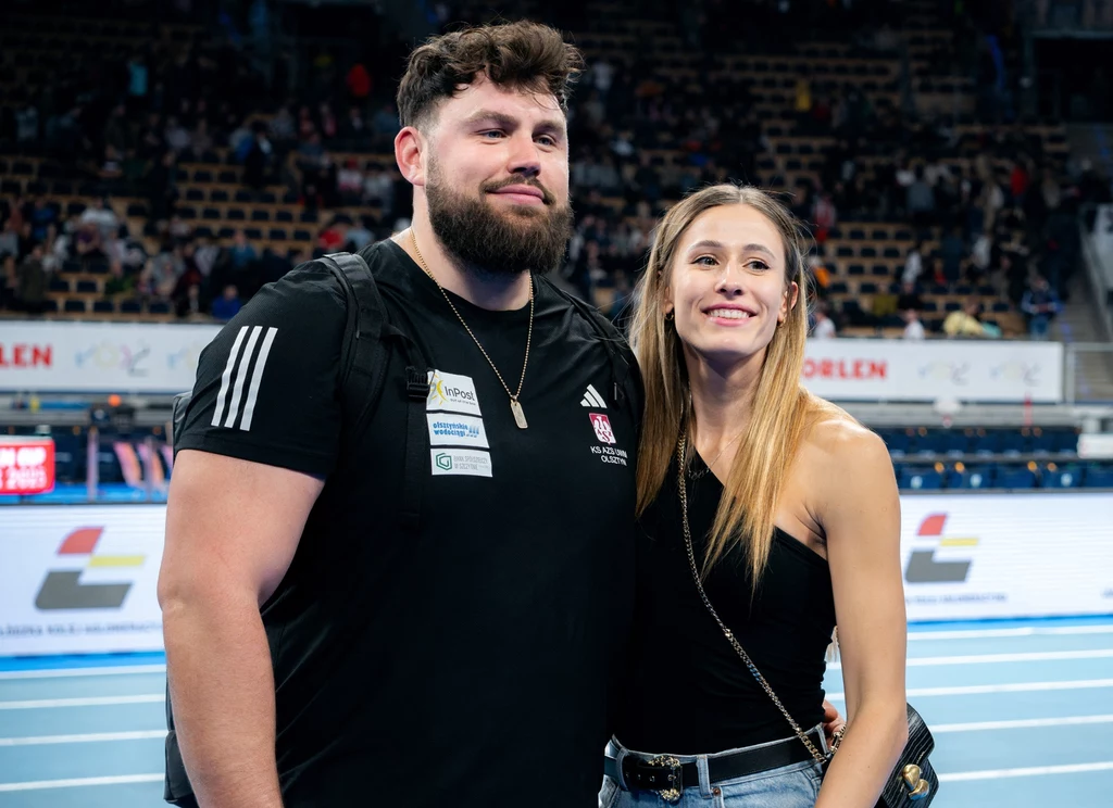 Dwoje uśmiechniętych młodych ludzi stoi razem na tle hali sportowej, mężczyzna ubrany w czarną koszulkę z logotypami, kobieta w czarnym topie i dżinsach, w tle trybuny z kibicami.