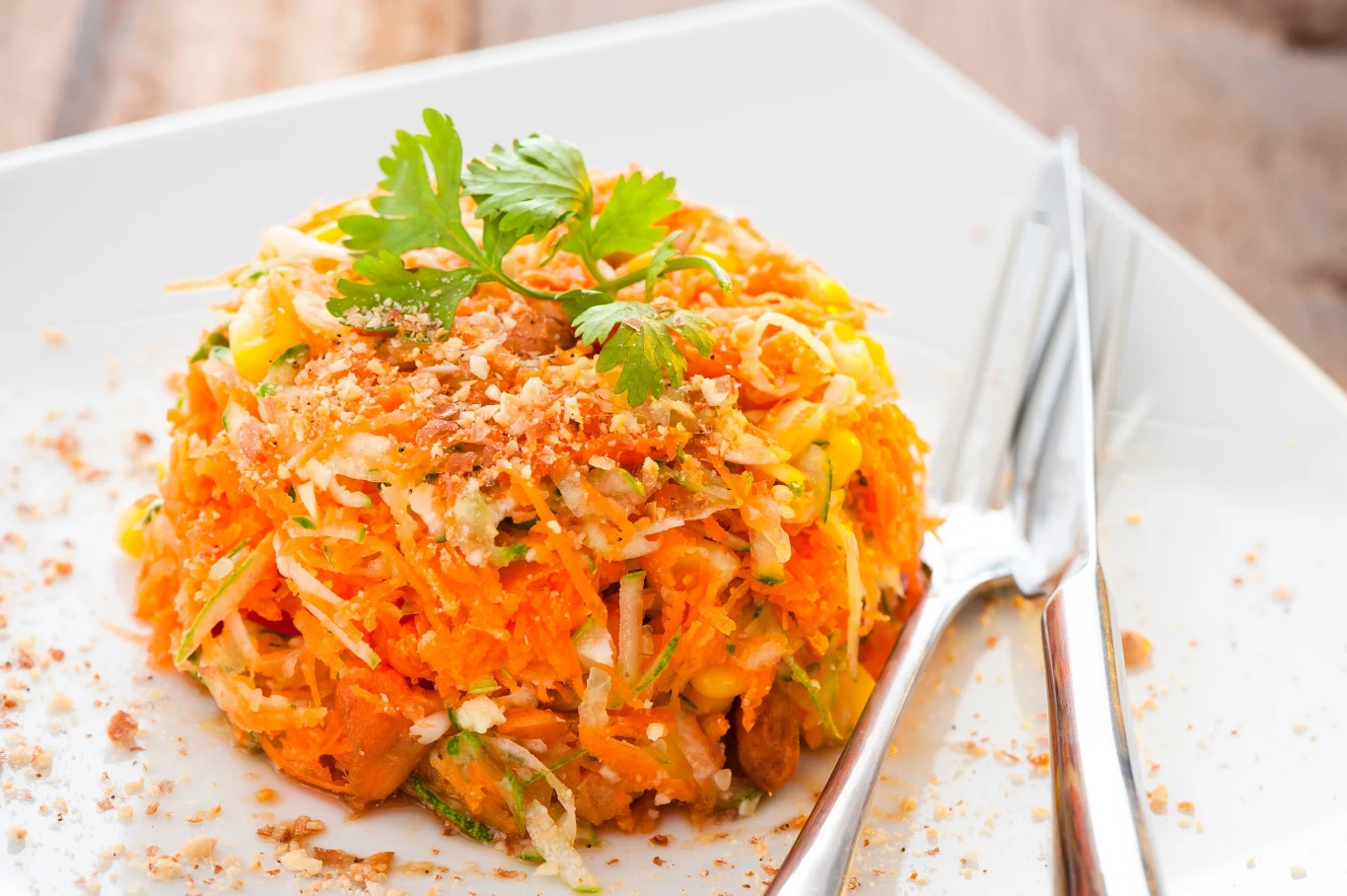 Marchew i kapusta pekińska to dobre połączenie Porcja świeżej sałatki z marchwi, posypanej kruszonymi orzechami i udekorowanej natką kolendry, podana na białym, kwadratowym talerzu z widelcem i nożem po prawej stronie.