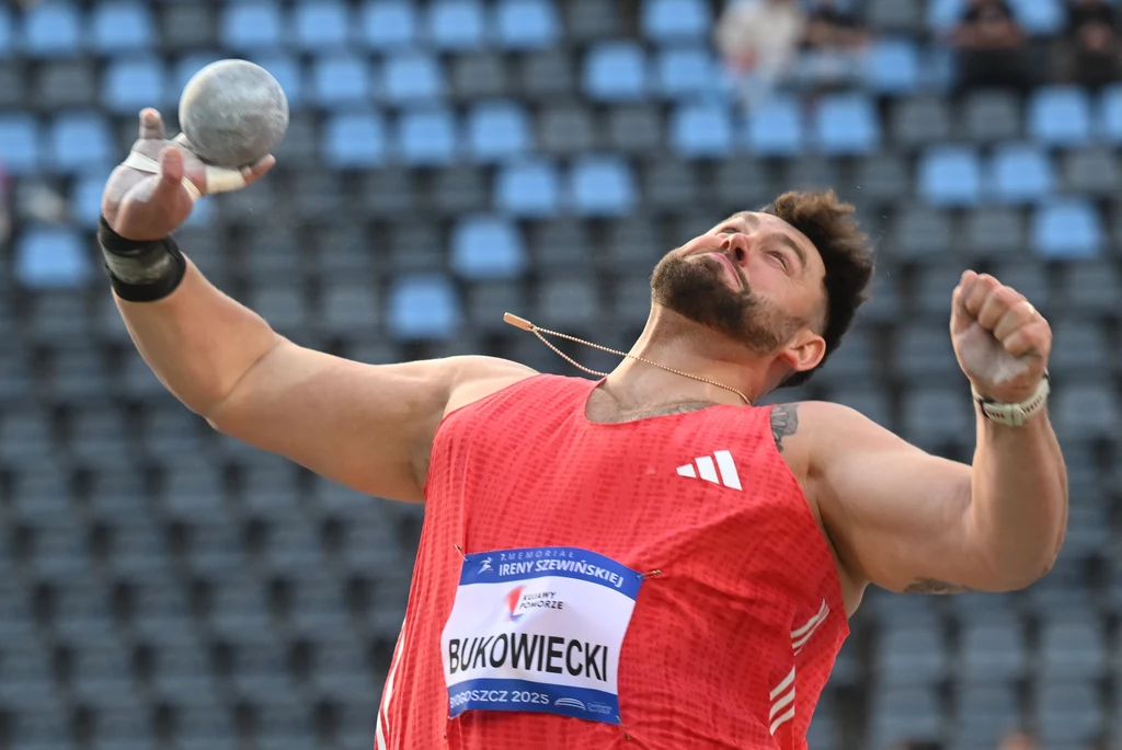 Konrad Bukowiecki Lekkoatleta w czerwonej koszulce podczas rzutu kulą na stadionie lekkoatletycznym, dynamiczna poza i skupienie na twarzy, w tle widoczne nieostre trybuny.