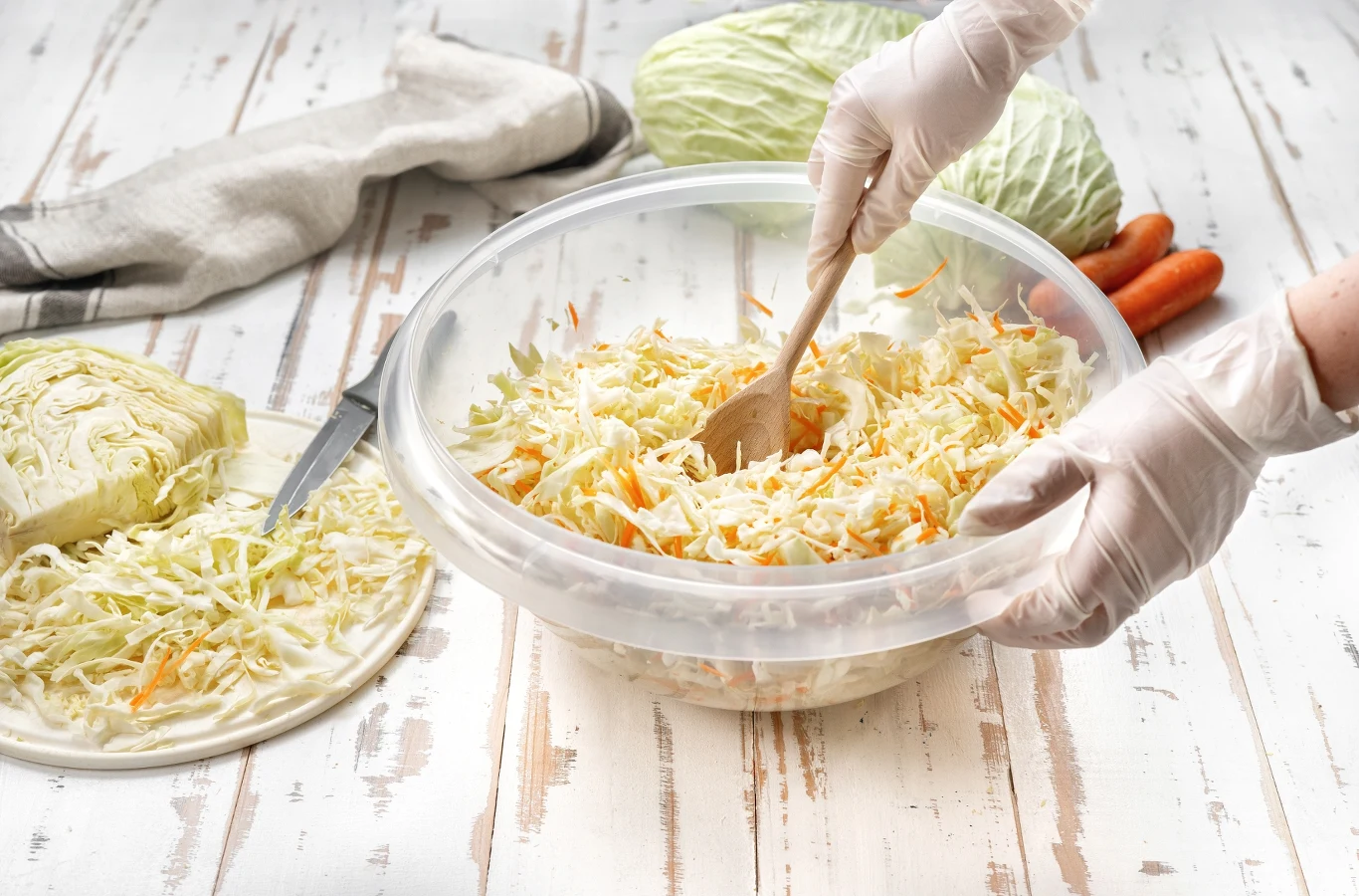 Colesław - warto przygotować większą porcję tej sałatki Ręce w jednorazowych rękawiczkach mieszają świeżo startą kapustę i marchewkę w dużej plastikowej misce, obok leży nóż oraz połowa główki kapusty i kilka marchewek, w tle znajduje się główka kapusty.