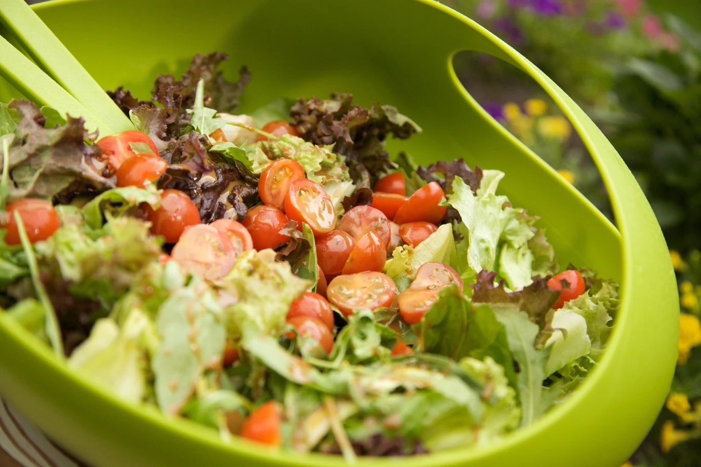 Sałatka z dressingiem figowym Świeża sałatka z mieszanki różnych rodzajów sałat i przekrojonych pomidorków koktajlowych w jasnozielonej misce, wszystko otoczone zielenią i kolorowymi kwiatami w tle.