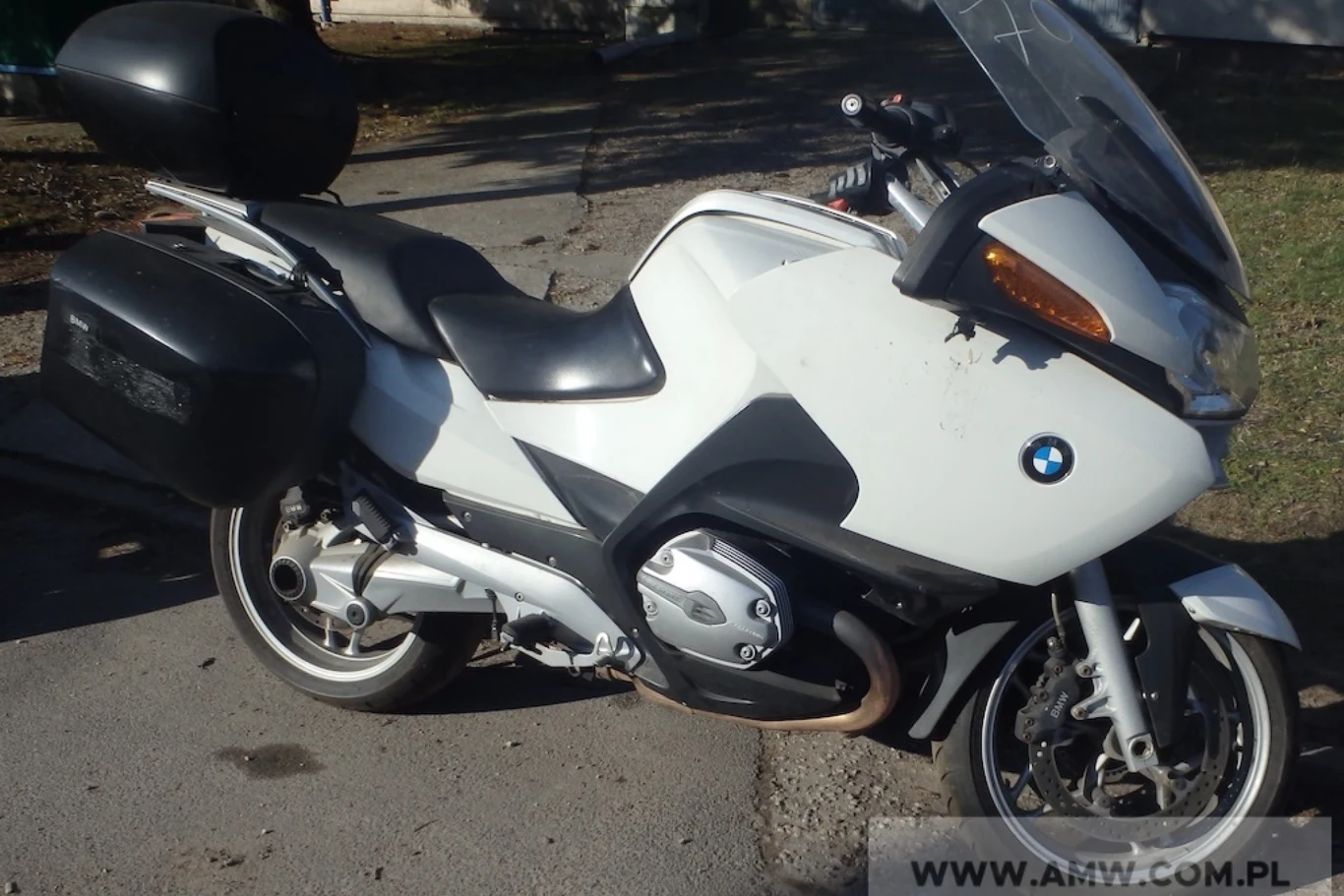 BMW R1200RT napędzany jest silnikiem typu bokser. AMW w Poznaniu wystawiła na sprzedaż łącznie cztery egzemplarze. Motocykl turystyczny marki BMW w kolorze białym, wyposażony w boczne kufry oraz tylni bagażnik, stoi zaparkowany na asfalcie przy krawężniku. W tle fragmenty trawnika i drzewa rzucające cień na pojazd.