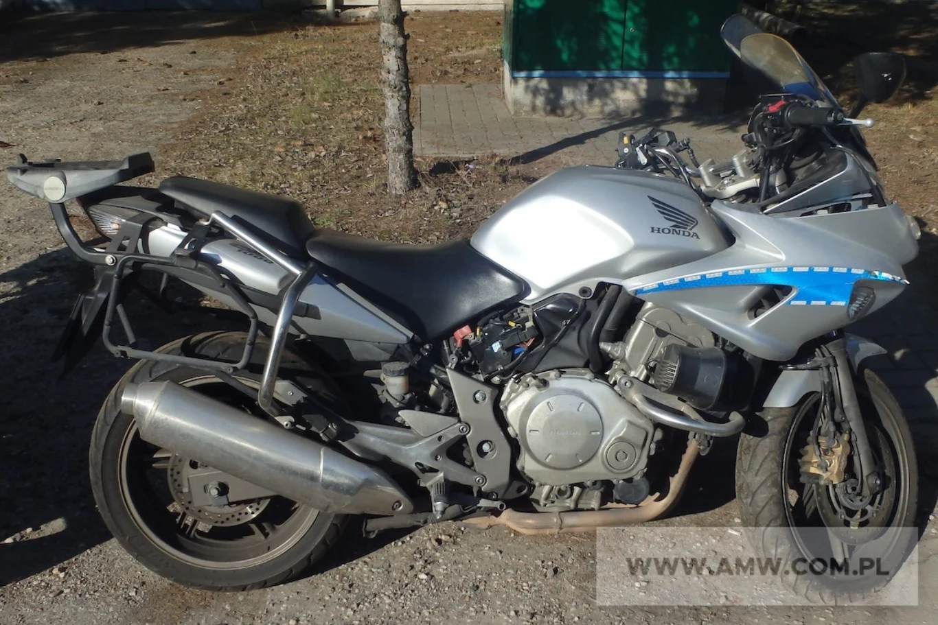 Honda CBF1000A przez lata stanowiła trzon polskiej drogówki. Srebrny motocykl Honda z niebieskimi akcentami, stojący na żwirowym podłożu, obok drzewa, z widocznym logo producenta oraz elementami wyposażenia typowymi dla pojazdów służbowych.