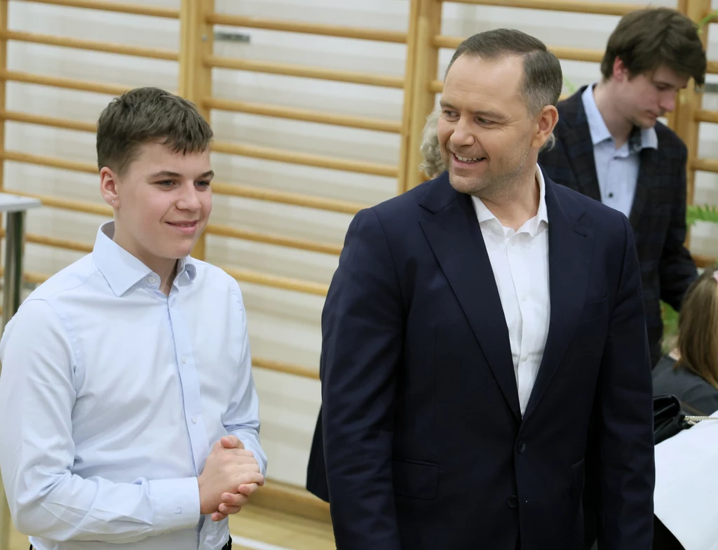 Karol Nawrocki z synem Dwójka mężczyzn ubranych elegancko rozmawia ze sobą w pomieszczeniu z drewnianymi drabinkami na ścianie, w tle znajdują się inne osoby zajęte rozmową lub pracą.