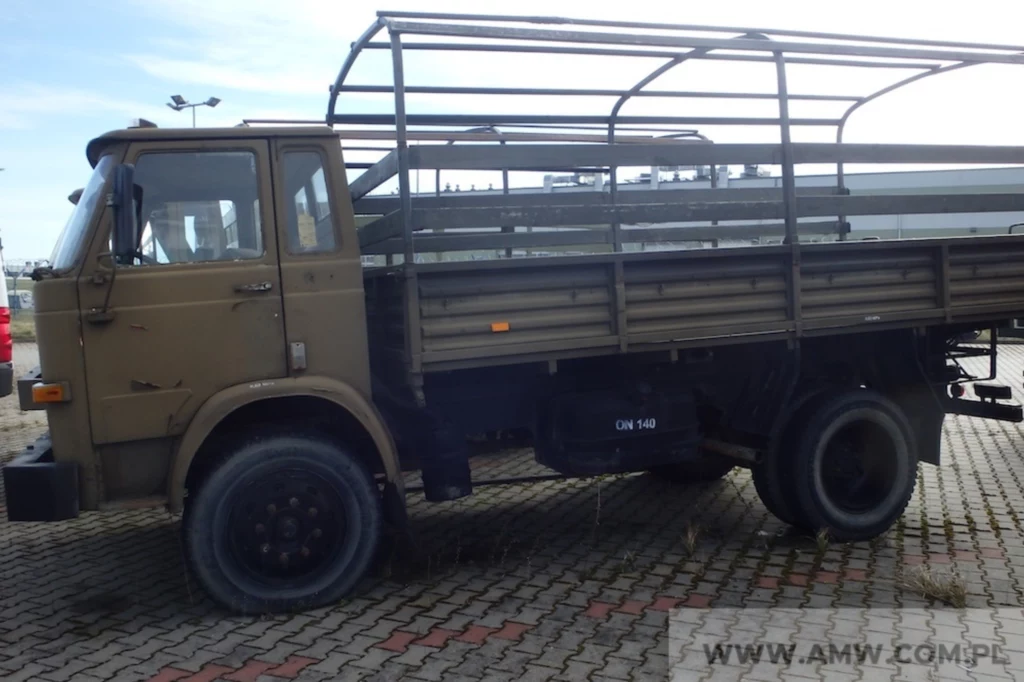 Star 200 produkowany był w Starachowicach w latach 1976-1994. Stara wojskowa ciężarówka w kolorze oliwkowym stoi na parkingu z nawierzchnią z kostki brukowej, nadwozie posiada otwartą ramę bez plandeki, a pojazd wykazuje ślady użytkowania.