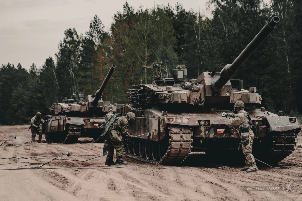 Czołgi K2GF na wyposażeniu Wojska Polskiego Dwa czołgi ustawione pośród leśnej drogi, wokół nich kilku żołnierzy w mundurach wykonuje czynności obsługowe, tło stanowią drzewa i ziemiste podłoże.