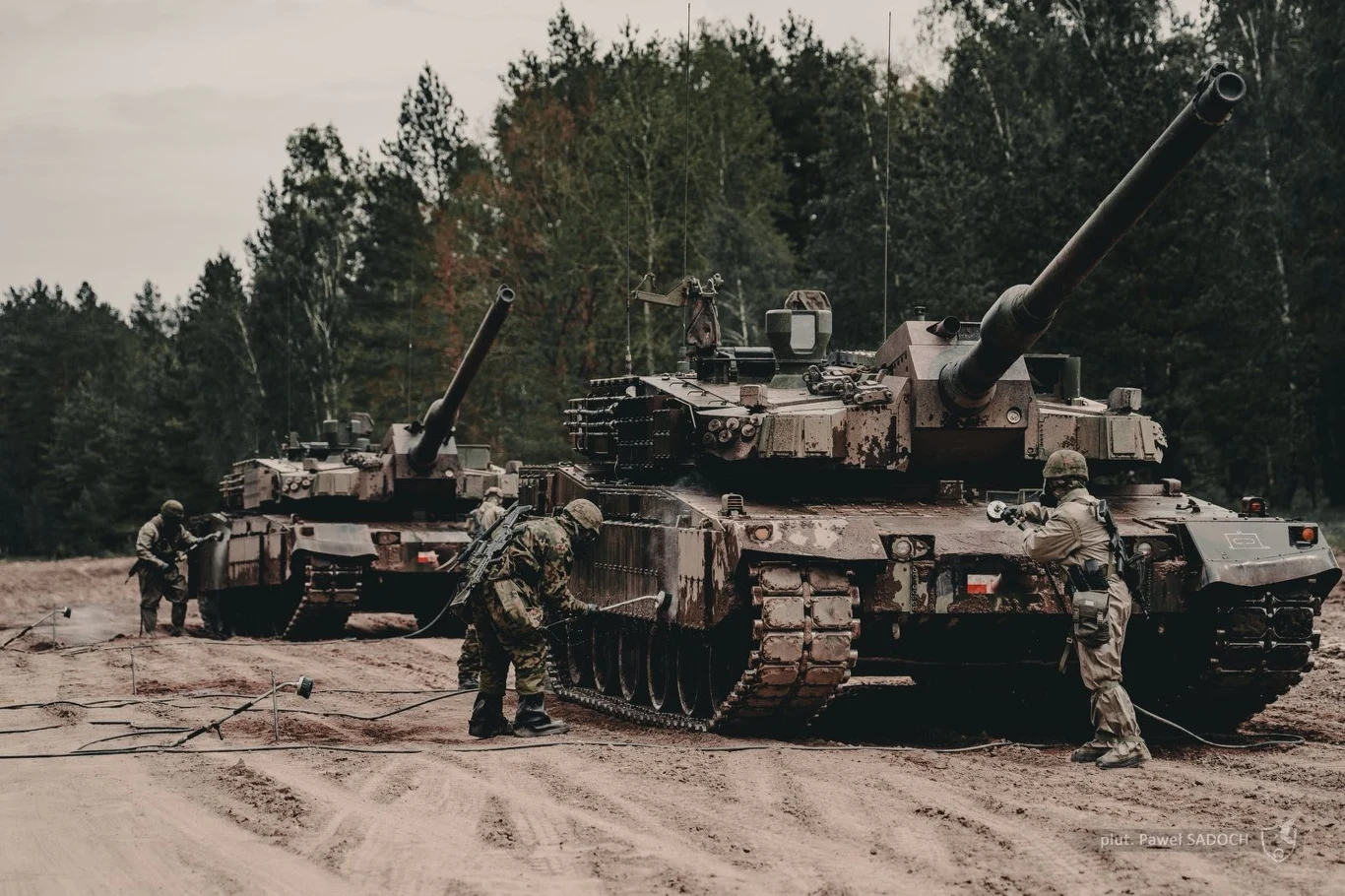 Dwa czołgi ustawione pośród leśnej drogi, wokół nich kilku żołnierzy w mundurach wykonuje czynności obsługowe, tło stanowią drzewa i ziemiste podłoże.