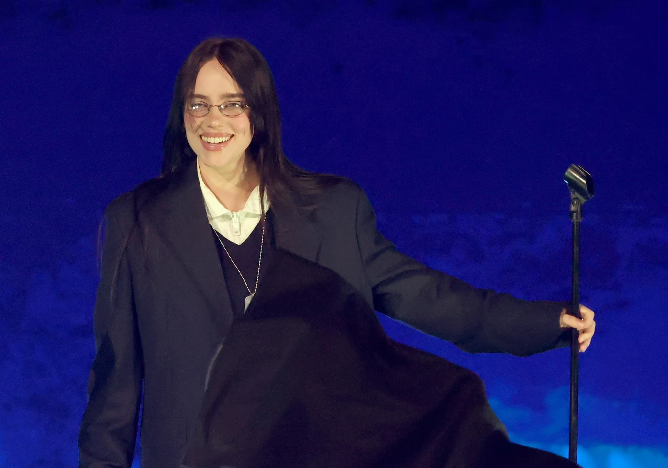 Głos Billie Eilish w krakowskim tramwaju! Uśmiechnięta osoba w okularach ubrana w ciemną, luźną marynarkę i białą koszulę stoi na scenie z mikrofonem na statywie, w tle widoczne niebieskie światło.