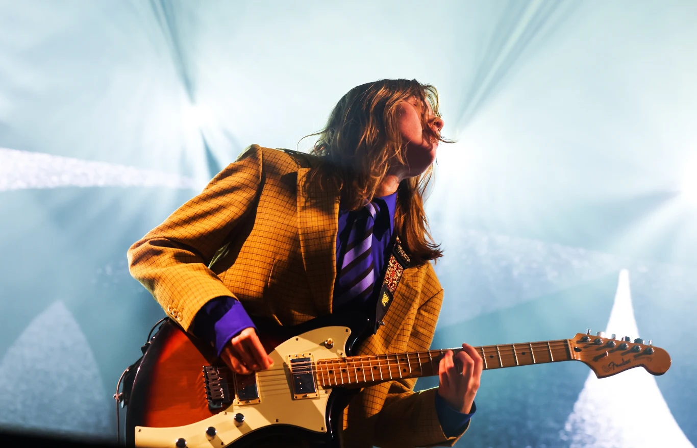 Girl in Red wystąpi na tegorocznej edycji Open'er Festivalu Młoda osoba w kolorowej marynarce i krawacie gra na gitarze elektrycznej na jasnym, podświetlonym tle sceny koncertowej, dynamiczna poza wskazuje na intensywny moment występu muzycznego.