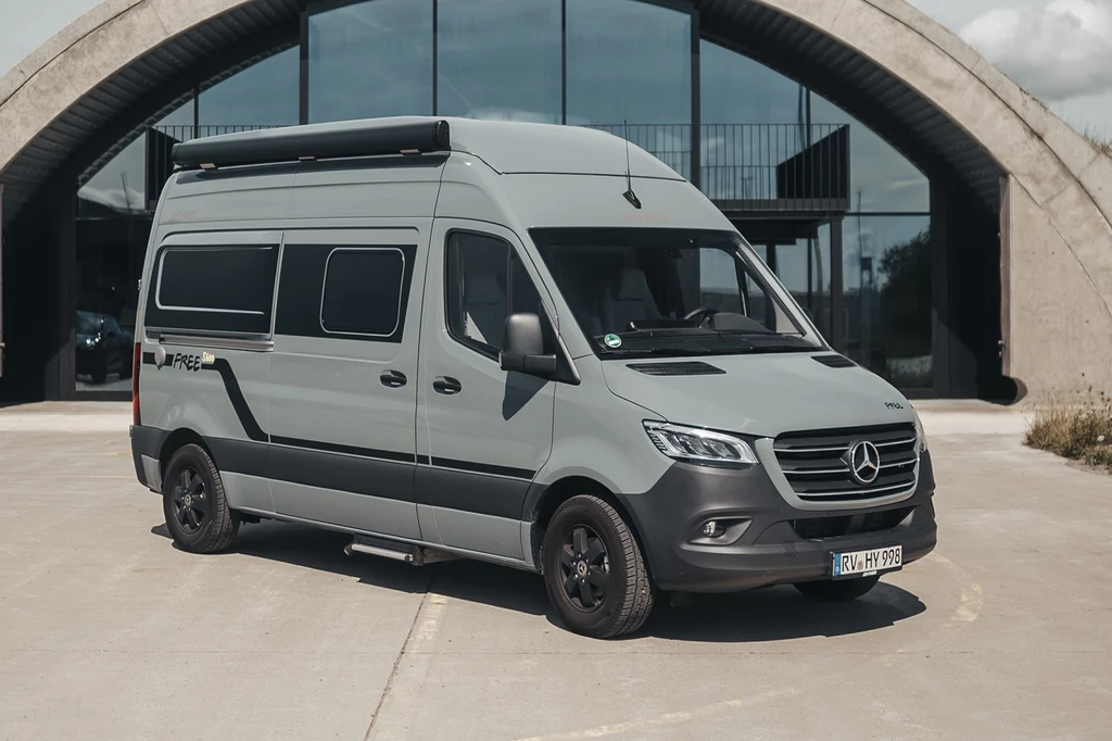 Andrzej Gut-Mostowy ma w kolekcji uterenowionego kampera . Srebrnoszary van Mercedes-Benz z zabudową kamperową zaparkowany na tle nowoczesnego budynku z dużymi przeszklonymi drzwiami i łukowatym wejściem.