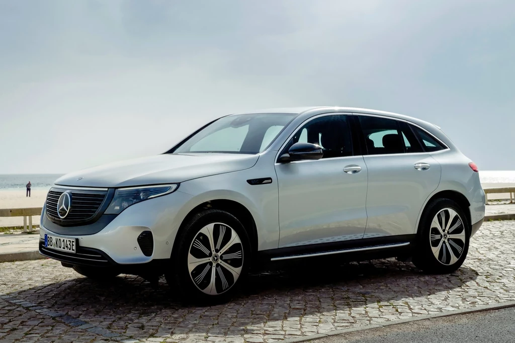 Mercedes EQC był pierwszym w historii modelem z gamy pojazdów elektrycznych niemieckiego producenta. Biały samochód osobowy marki Mercedes-Benz zaparkowany na brukowanej drodze przy nabrzeżu, w tle widoczny fragment morza i lekko zachmurzone niebo.