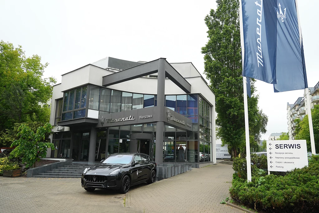 Otwarcie salonu w Warszawie wpisuje się w szerszą strategię inwestycyjną Maserati. Nowoczesny budynek salonu Maserati z dużymi przeszkleniami, przed wejściem zaparkowany luksusowy samochód tej marki, po prawej stronie widoczne logo i flaga Maserati oraz tablica informacyjna serwisu, wokół zieleń i drzewa.