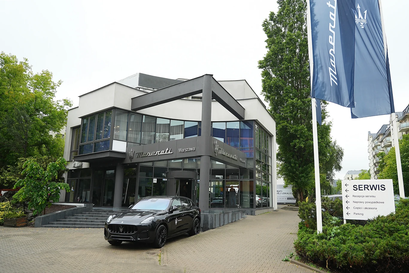 Nowoczesny budynek salonu Maserati z dużymi przeszkleniami, przed wejściem zaparkowany luksusowy samochód tej marki, po prawej stronie widoczne logo i flaga Maserati oraz tablica informacyjna serwisu, wokół zieleń i drzewa.