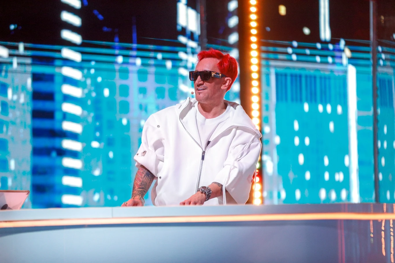 Michał Wiśniewski będzie też gościem Akademii Sztuk Przepięknych podczas Pol'and'Rock Festivalu Mężczyzna z czerwonymi włosami i tatuażami, ubrany w białą kurtkę, stojący za futurystycznym biurkiem w jasnym świetle na tle nowoczesnej, podświetlonej scenografii, noszący ciemne okulary przeciwsłoneczne.