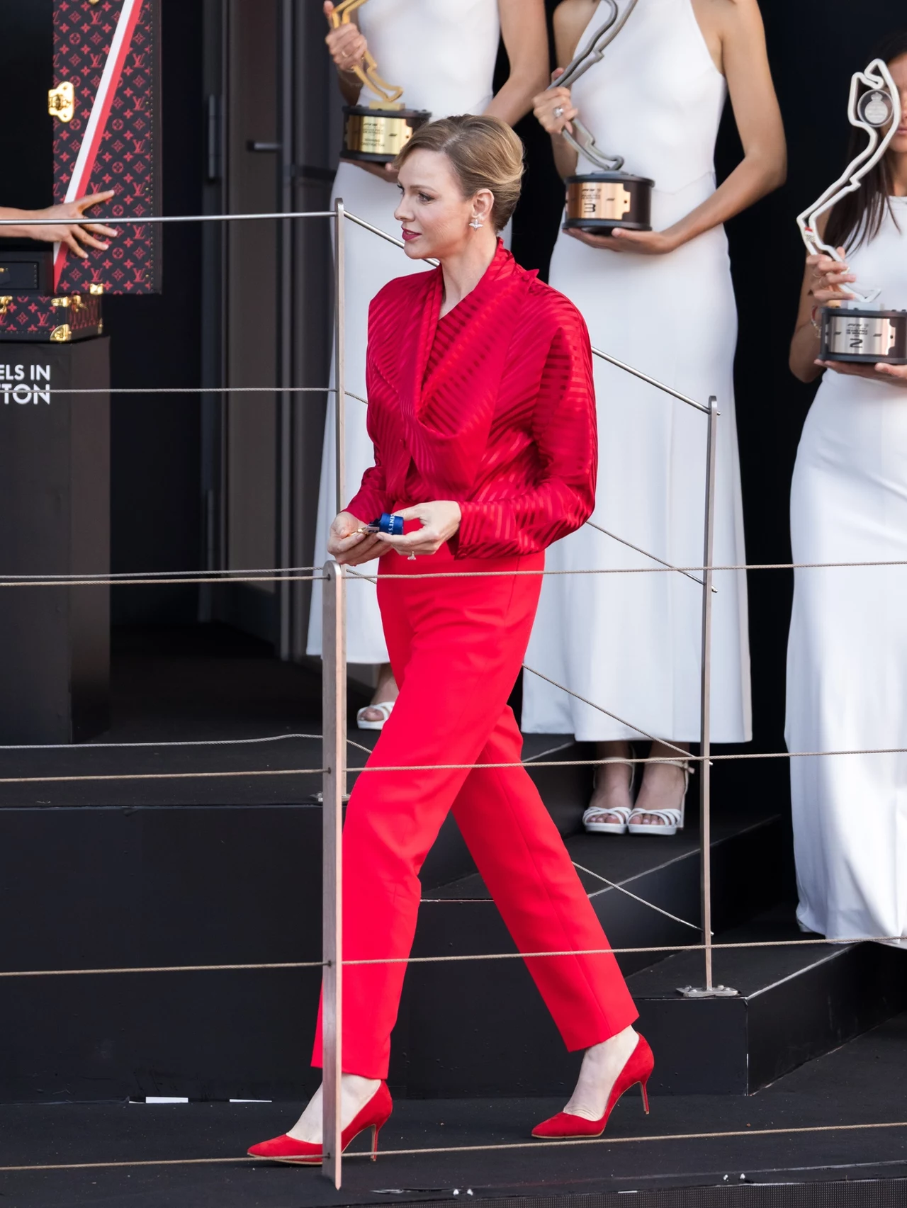 Księżna Charlene postawiła na mocny akcent kolorystyczny Elegancko ubrana kobieta w czerwonym garniturze i szpilkach przechodzi obok osób w białych sukniach trzymających statuetki, scena ma charakter oficjalny i uroczysty.
