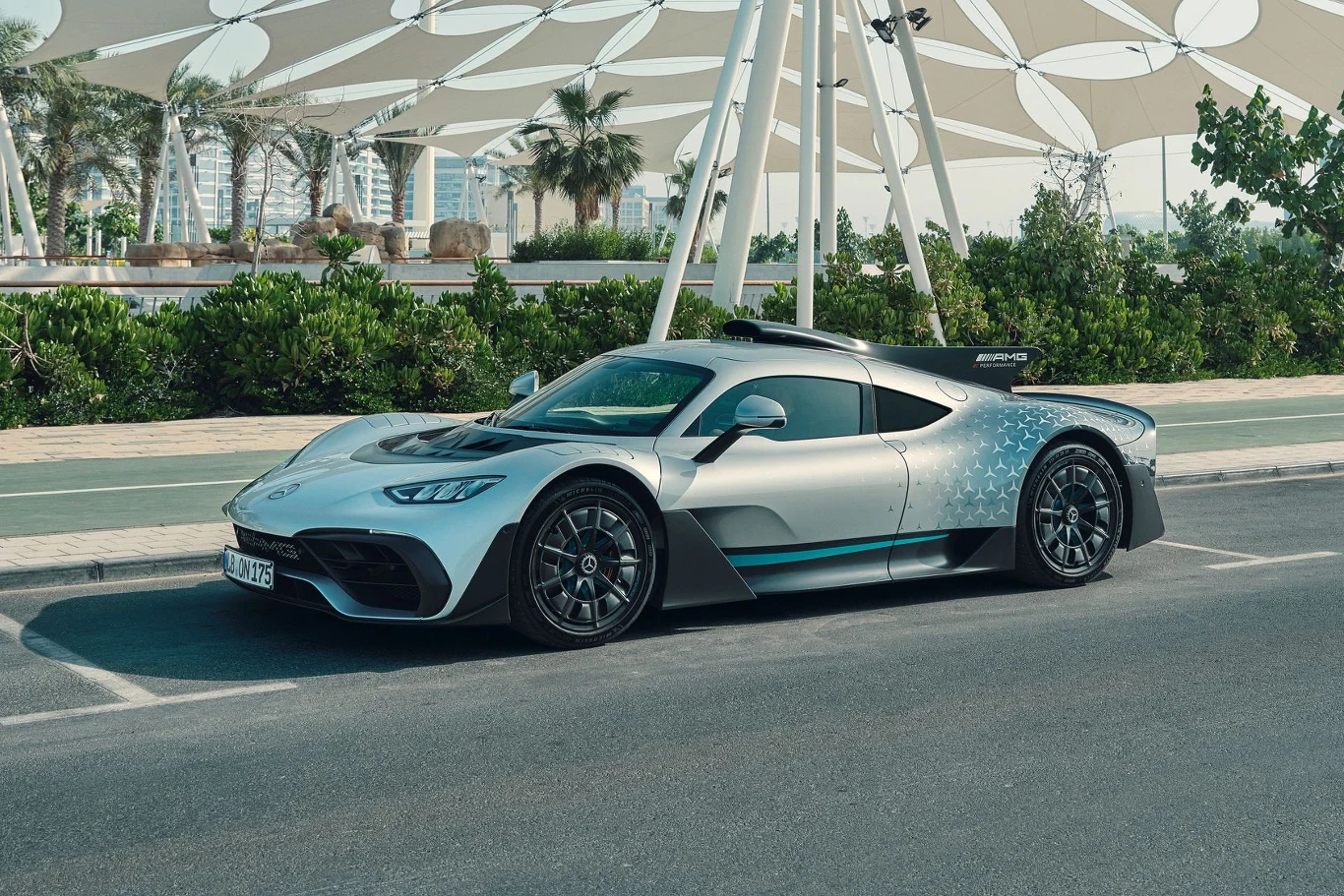 Mercedes-AMG One to właściwie bolid F1 z innym nadwoziem i homologacją drogową Srebrny supersamochód Mercedes-AMG One zaparkowany na ulicy, w tle nowoczesna architektura z białymi strukturami i palmami.