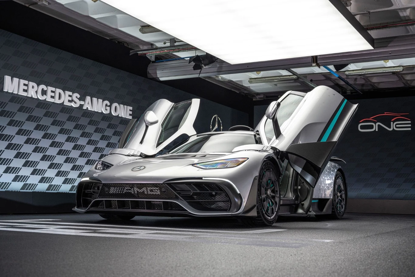 Już tylko 273 sztuki Mercedesa-AMG One jeżdżą po świecie. Srebrny supersamochód Mercedes-AMG One z otwartymi do góry drzwiami, ustawiony w nowoczesnym, oświetlonym salonie ekspozycyjnym, z wyeksponowanym logo marki na tle ściany z napisem ONE.
