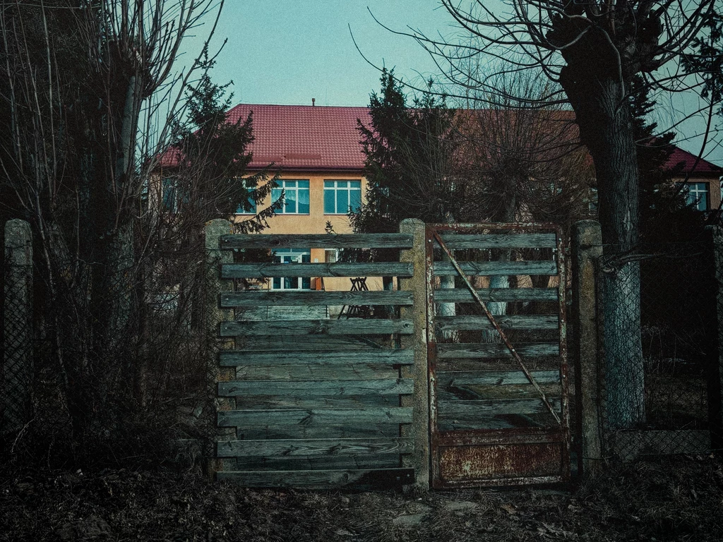 Dom opieki Jesień Podlasia Zniszczona drewniana brama prowadząca na teren otoczony starym ogrodzeniem z siatki; za bramą widoczny żółty budynek z czerwonym dachem, przesłonięty częściowo przez drzewa bez liści.