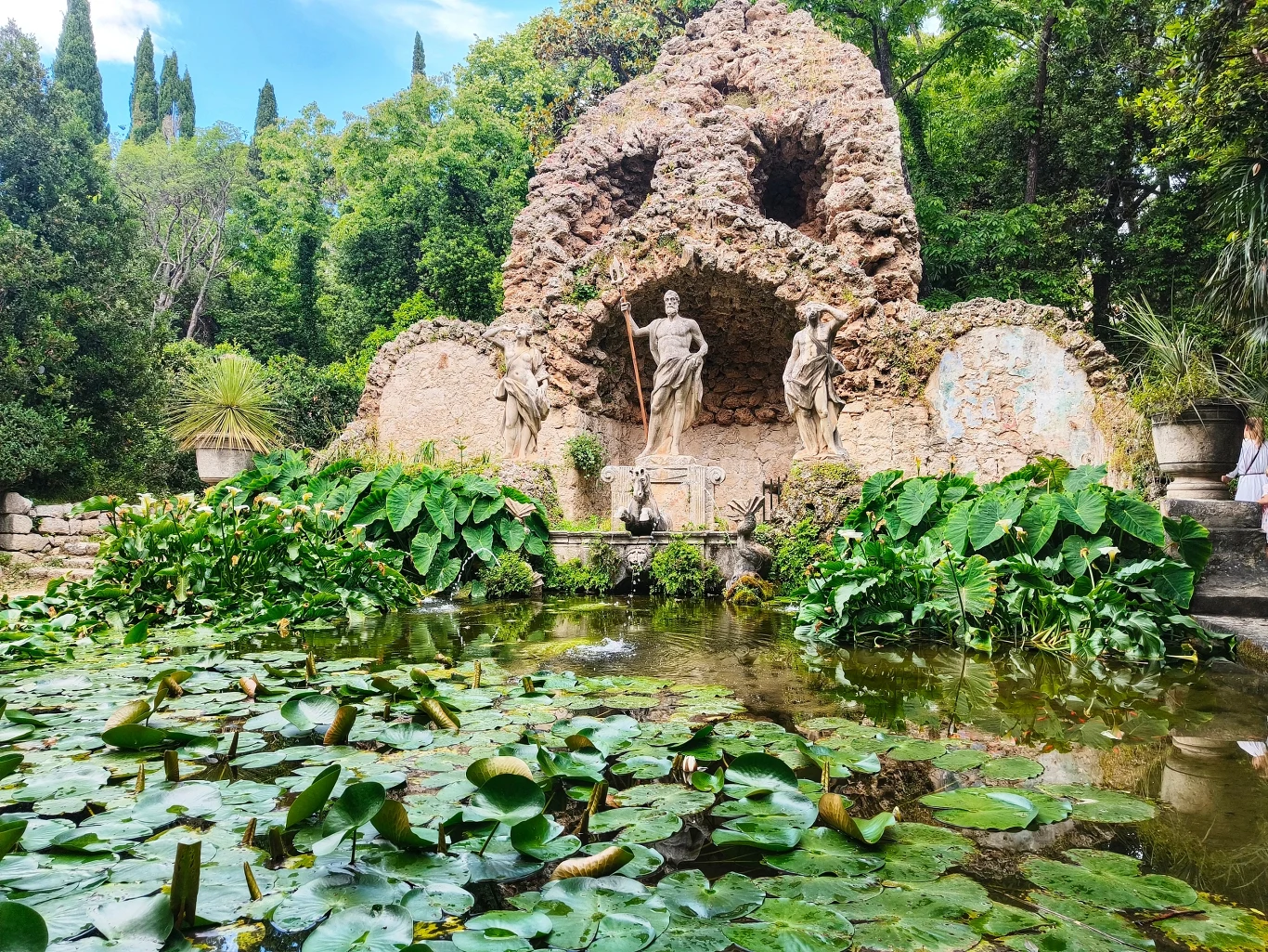 Kamienna grota ozdobiona rzeźbami postaci mitologicznych, otoczona gęstą zielenią i małym stawem porośniętym liliami wodnymi, w tle widoczne wysokie drzewa.
