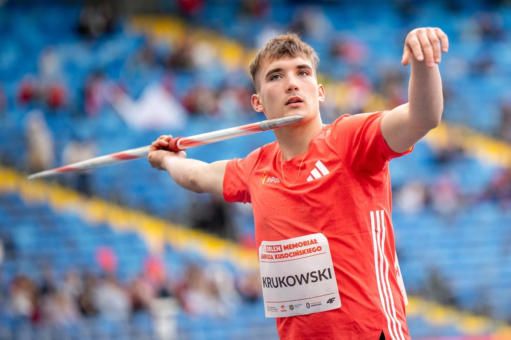 Roch Krukowski Młody sportowiec w czerwonej koszulce przygotowuje się do rzutu oszczepem na stadionie z niebieskimi trybunami w tle, skupiony na technice i skoncentrowany na swojej konkurencji.