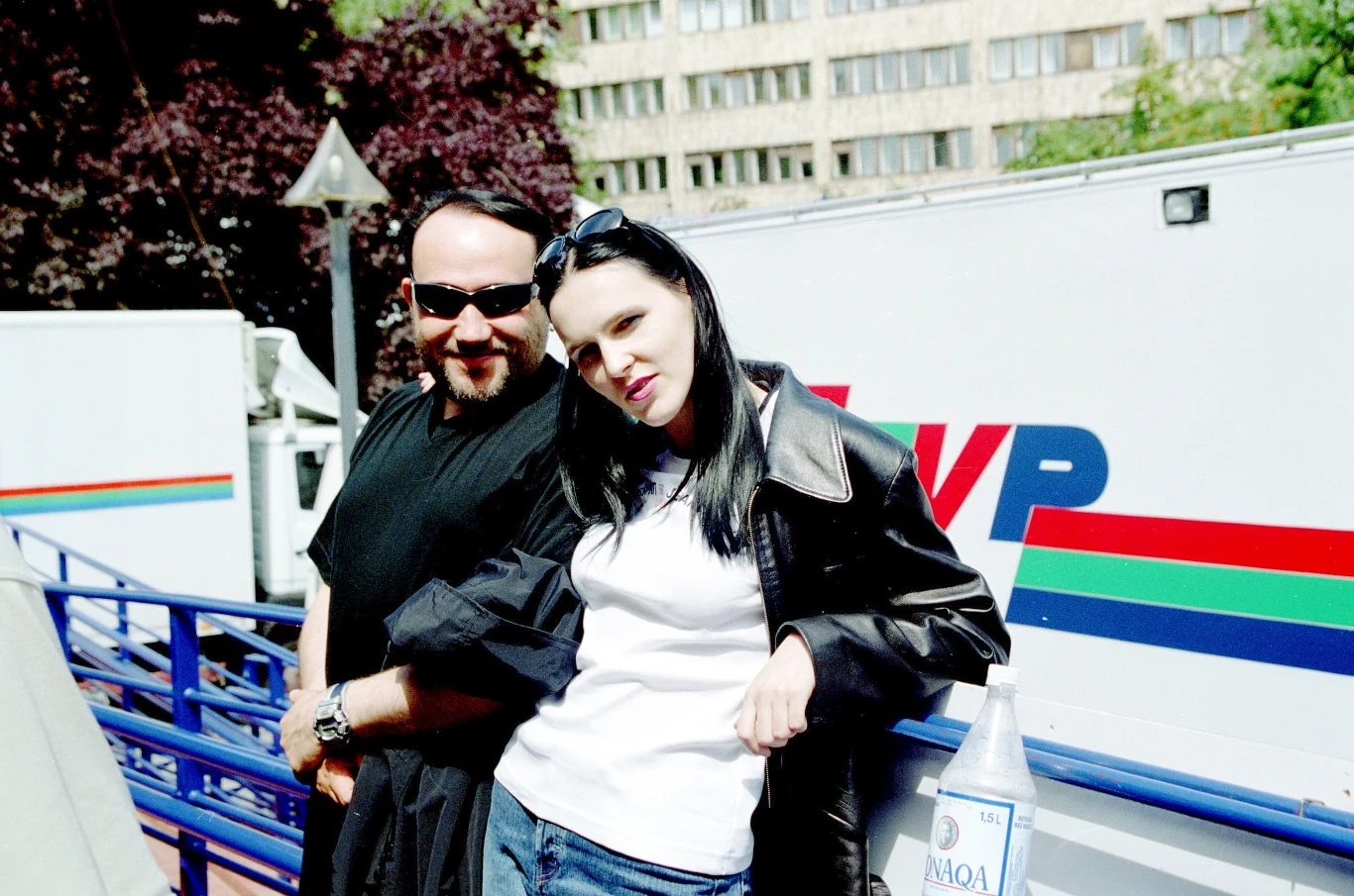 Grzegorz Skawiński i Agnieszka Chylińska (O.N.A.) w 1999 r. Dwie osoby stoją oparte o niebieską barierkę na tle samochodu z logo TVP, mężczyzna ubrany jest na czarno, kobieta w białej koszulce i czarnej skórzanej kurtce, obok butelka wody mineralnej.