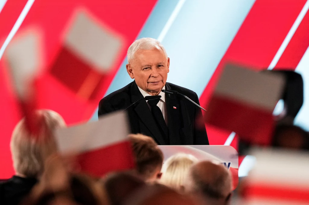 Jarosław Kaczyński podczas wieczoru wyborczego po drugiej turze wyborów prezydenckich 2025 Starszy mężczyzna stojący przy mównicy podczas wydarzenia politycznego, w tle widoczne są polskie flagi i rozmyte sylwetki ludzi. Główne elementy sceny to poważny wyraz twarzy mężczyzny i patriotyczna atmosfera zdominowana narodowymi barwami.