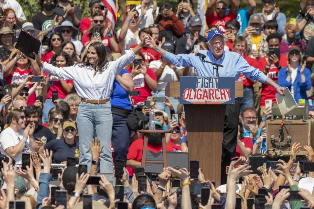 Alexandria Occasio-Cortez i Bernie Sanders na wiecu w Los Angeles Tłum ludzi zgromadzony podczas wiecu politycznego na otwartym powietrzu, dwie główne postacie stoją razem na scenie, podnosząc ręce do góry, na mównicy widoczny napis FIGHT OLIGARCHY, ludzie w tłumie unoszą ręce i nagrywają wydarzenie telefonami.
