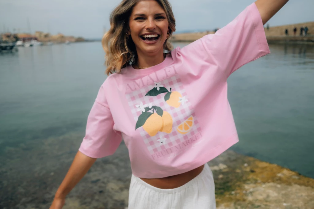 Lekkie, oddychające tkaniny, luźne kroje, falbanki, pastele, zabawne włoskie grafiki Uśmiechnięta młoda kobieta z blond włosami ubrana w różowy t-shirt z nadrukiem cytryn oraz białą spódnicę stoi przy brzegu wody, z rozłożonymi ramionami, na tle portu lub przystani.