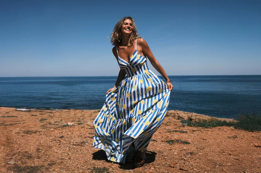 Ubrania z letniej kolekcji NAREE są delikatne i eleganckie Kobieta w długiej, kolorowej sukience stoi na plaży, uśmiechnięta, z rozwianymi włosami na tle morza i czystego nieba.