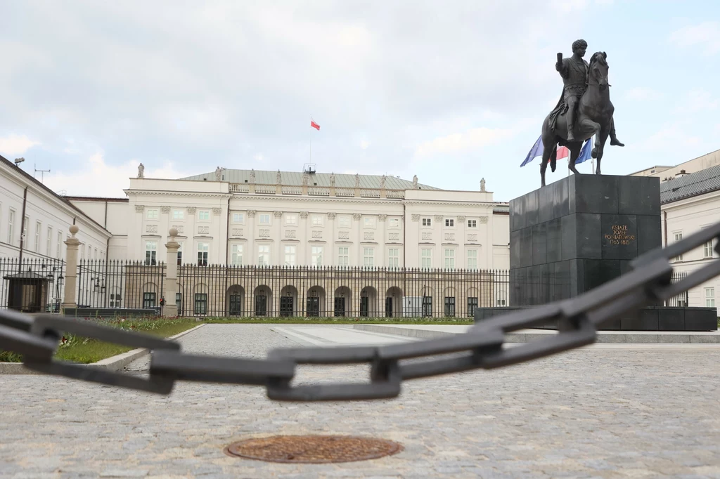 Pałac Prezydencki w Warszawie Pałac z wysokim ogrodzeniem oraz łańcuchem oddzielającym od wejścia, na pierwszym planie widoczny pomnik jeźdźca na koniu na wysokim postumencie, polska flaga powiewa na dachu budynku.