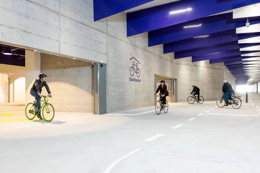 Nowy tunel w Zurychu spodobał się rowerzystom. Zabiegali o niego od 14 lat Ludzie jadący rowerami w nowoczesnym, zadaszonym parkingu rowerowym o betonowych ścianach i charakterystycznym niebieskim suficie.