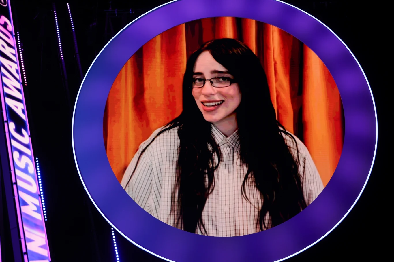 Billie Eilish da dwa koncerty w Krakowie Młoda kobieta o długich, ciemnych włosach i okularach, ubrana w jasną, kraciastą koszulę, uśmiecha się na tle pomarańczowej zasłony, widoczna w dużym okrągłym kadrze oprawionym fioletową ramką.