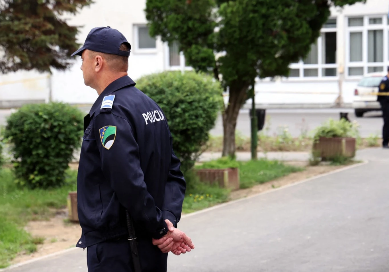 Władze i służby w bośniackim mieście walczą ze stanem klęski żywiołowej . Zdj. ilustracyjne Policjant w mundurze patroluje teren w pobliżu budynku, wokół zieleń i krzewy, w tle widoczna kolejna osoba w mundurze.