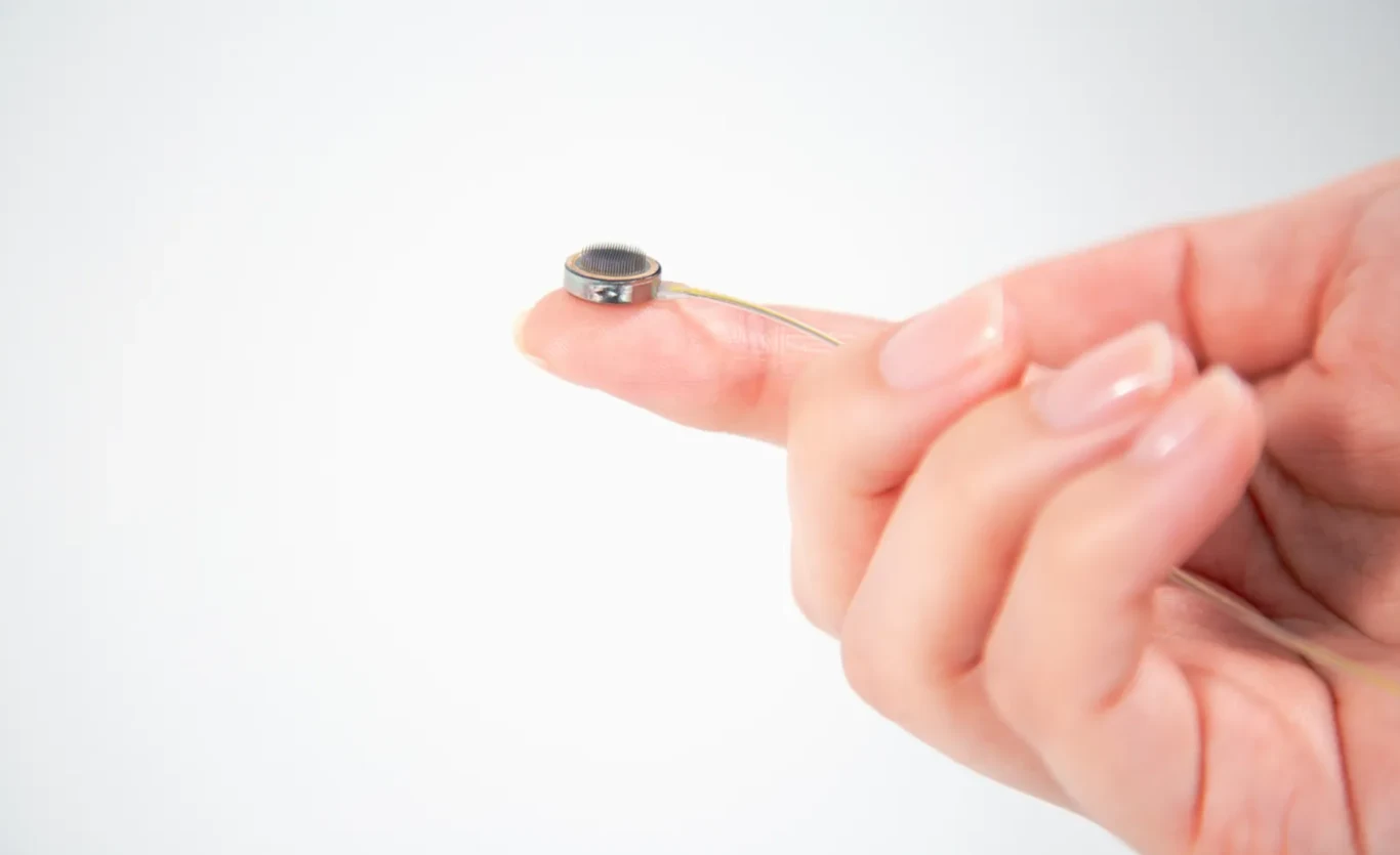 Paradromics wszczepia pierwszy interfejs mózg-komputer człowiekowi. Wielki krok w neurotechnologii Dłoń trzymająca mały mikrofon elektretowy na opuszkach palców, z wyraźnie widocznymi cienkimi przewodami wystającymi z urządzenia, białe tło, skupienie na szczegółach mikrofonu.