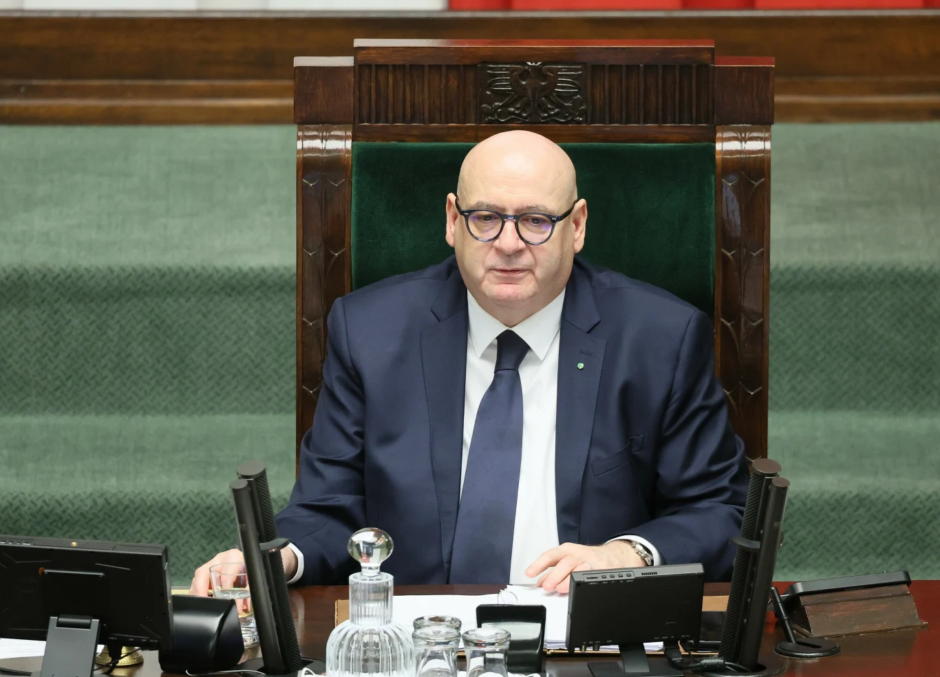 Wicemarszałek Sejmu Piotr Zgorzelski Mężczyzna w okularach i garniturze siedzący za dużym, drewnianym biurkiem, tło stanowią zielony dywan i fragment paneli ściennych, wokół widoczne są mikrofony, dokumenty i szklany dzbanek z wodą.