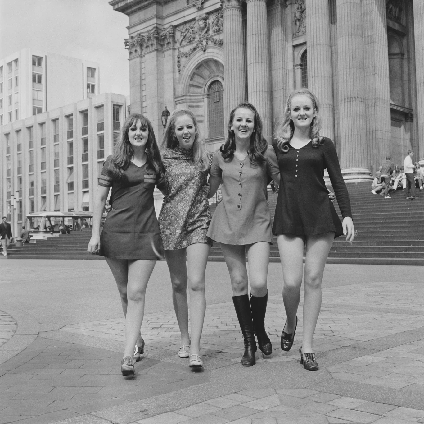 Marcie Jones (po lewej) z zespołem Marcie and The Cookies Cztery młode kobiety ubrane w krótkie sukienki idą razem ulicą w mieście, w tle monumentalna architektura oraz schody prowadzące do dużego budynku.