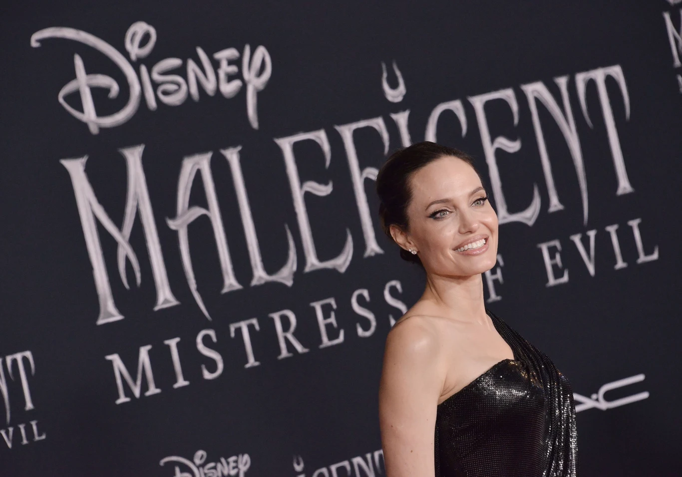 Kobieta w czarnej, eleganckiej sukni z odkrytym ramieniem stoi przed ścianą z logo Disney i tytułem filmu Maleficent Mistress of Evil, uśmiecha się szeroko, tło ciemne z dużymi, białymi napisami.