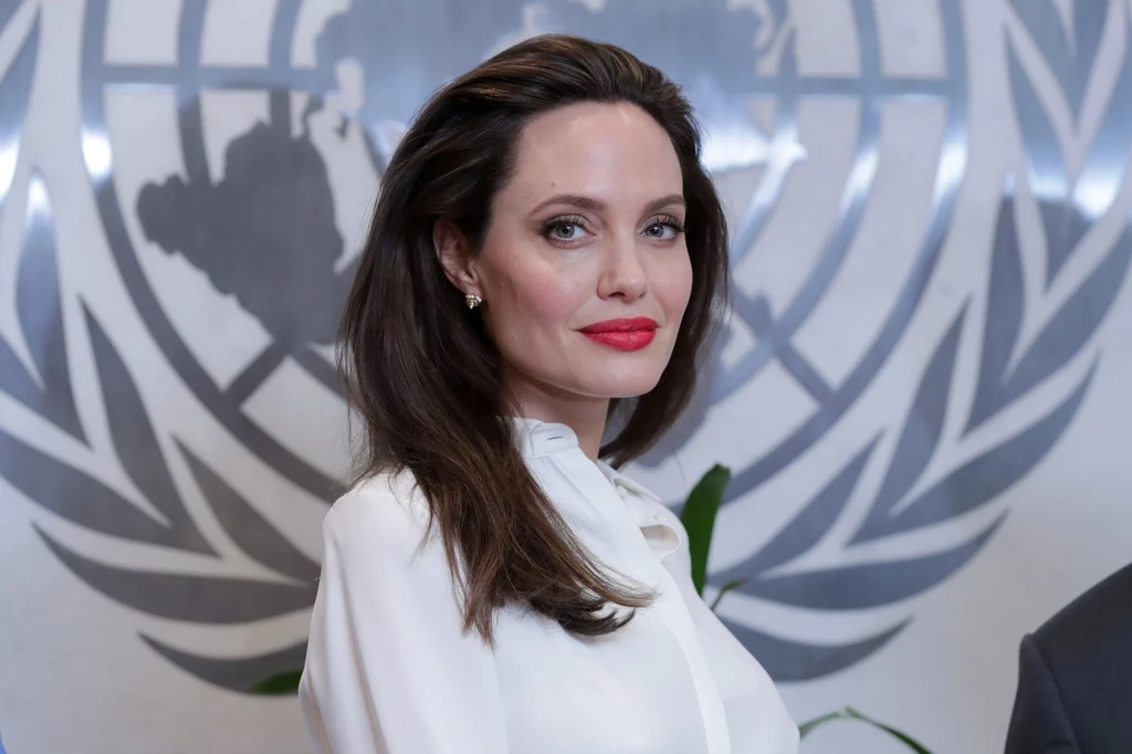 Angelina Jolie stanowi również wzór dla swoich dzieci, które wychowywane są przez nią na empatycznych ludzi Elegancka kobieta z długimi brązowymi włosami ubrana w białą bluzkę stoi na tle logo ONZ, jej wyraz twarzy jest poważny a makijaż podkreśla czerwone usta.