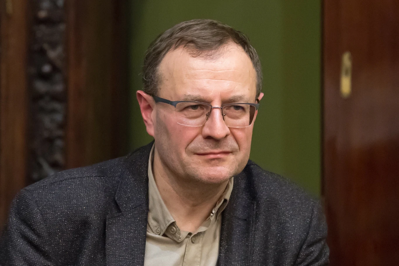 Wybory prezydenckie 2025. Prof. Antoni Dudek komentuje wyniki wyborów Mężczyzna w średnim wieku w okularach, ubrany w ciemną marynarkę i jasną koszulę, siedzi na tle zielonej ściany oraz drewnianych drzwi.