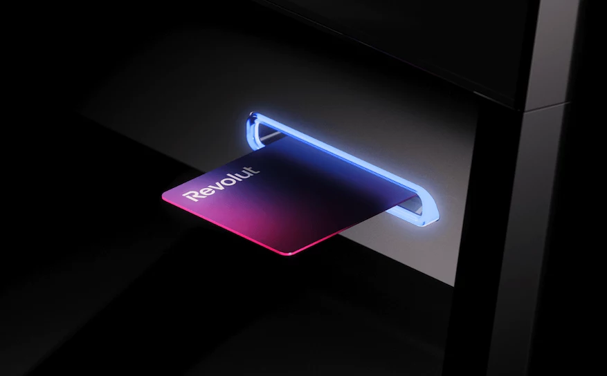 Karta płatnicza Revolut wsuwana do podświetlonego czytnika lub bankomatu, mocny kontrast kolorów i nowoczesne oświetlenie podkreślają technologiczny charakter sceny.