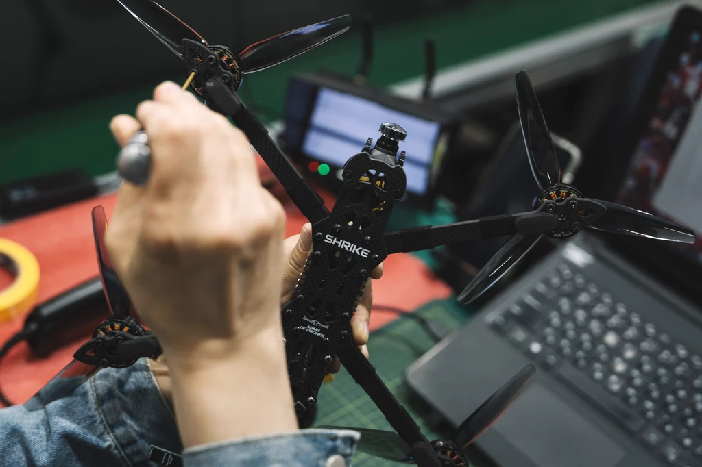 Produkcja ukraińskich dronów Shrike FPV firmy Skyfall Ręka osoby dokonująca naprawy lub montażu drona wyścigowego z napisem 'SHRIKE', w tle widoczna klawiatura laptopa i narzędzia.
