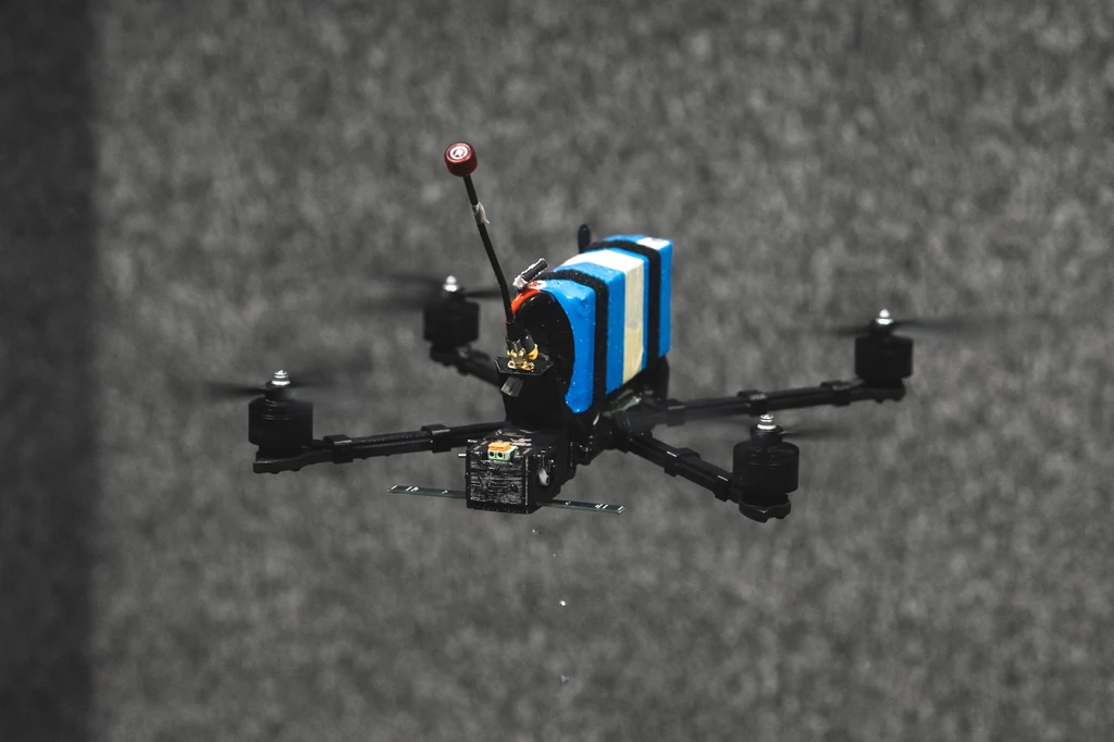 Lot testowy drona typu Shrike FPV Czterowirnikowy dron z zamontowaną elektroniką oraz niebiesko-białym elementem transportowym w centrum kadru na rozmytym, szarym tle.