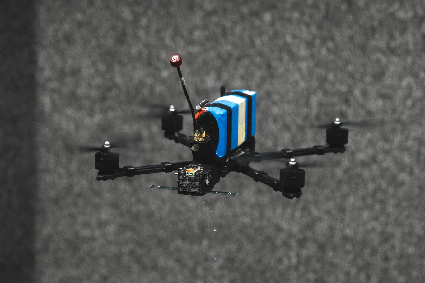 Czterowirnikowy dron z zamontowaną elektroniką oraz niebiesko-białym elementem transportowym w centrum kadru na rozmytym, szarym tle.