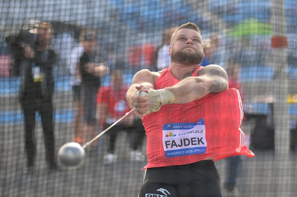 zawodnik wykonujący rzut młotem na stadionie lekkoatletycznym, ujęcie dynamiczne, młot w połowie obrotu, siatka ochronna w tle, publiczność w rozmytym tle
