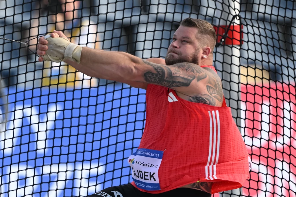 Sportowiec w czerwonej koszulce wykonuje rzut młotem podczas zawodów lekkoatletycznych, widoczny jest w klatce rzutowej z napiętą linką do młota; tło stanowią trybuny i reklamy.